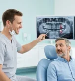 A male dentist in light gray scrubs explaining a dental X-ray, comparing root canal treatment vs tooth extraction, to a male patient.