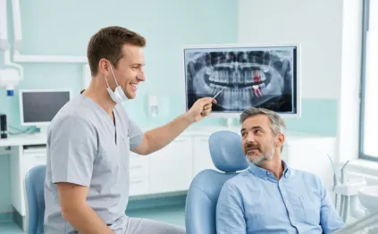 A male dentist in light gray scrubs explaining a dental X-ray, comparing root canal treatment vs tooth extraction, to a male patient.