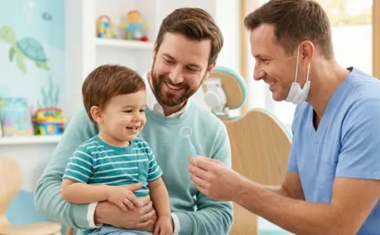 A friendly male paediatric dentist Muscat showing a small dental mirror to a happy toddler sitting on his father's lap.